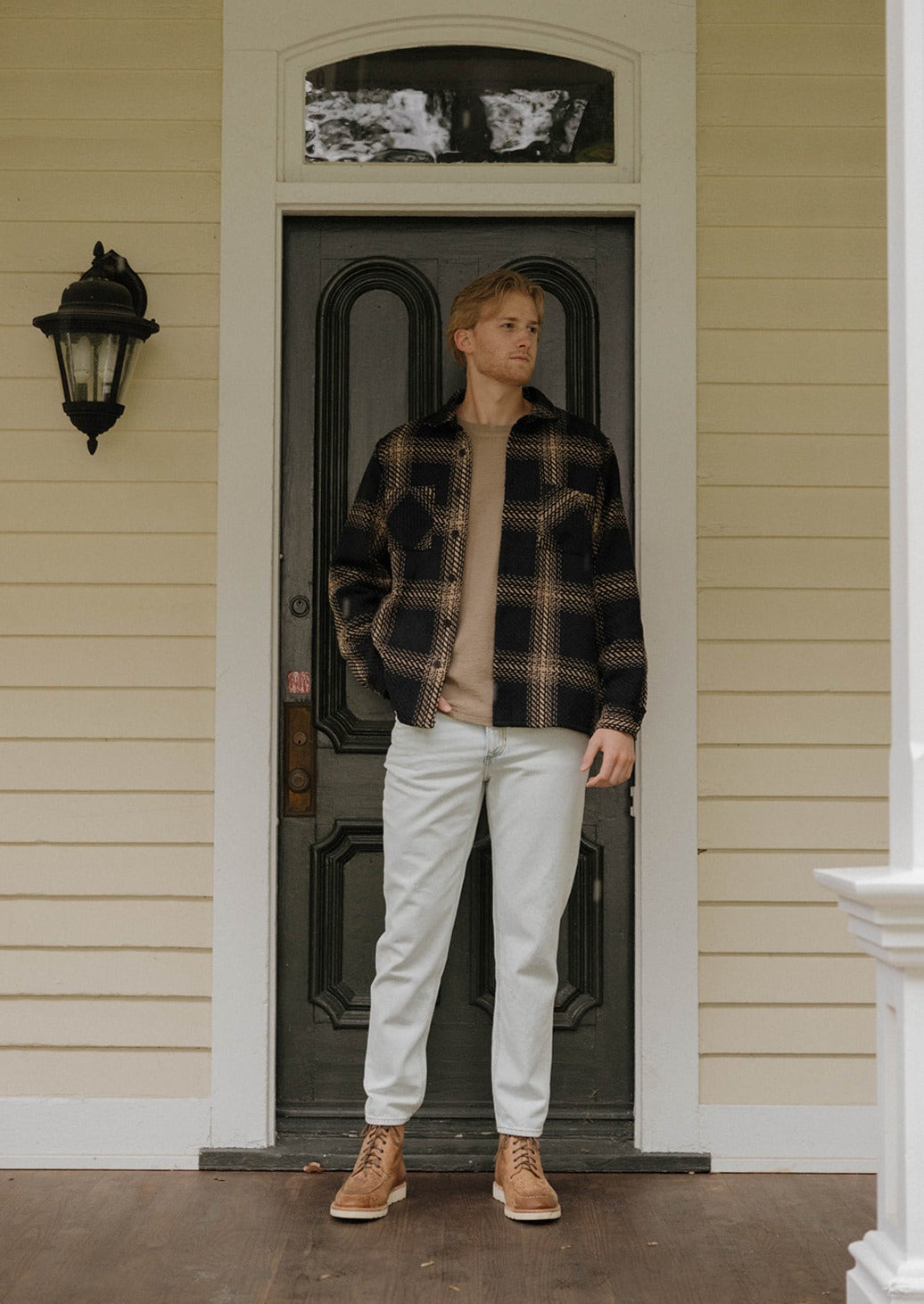 A male model is standing in front of a door of a house while wearing the Joybird Carrot Jeans styled with a black and tan plaid shacket