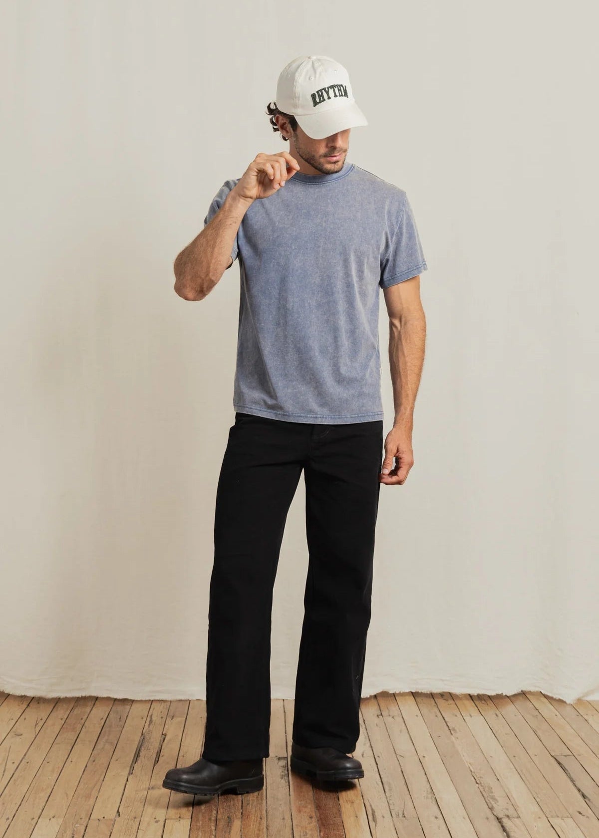 Person wearing a gray t-shirt, black pants, and a white cap with text on a wooden floor.