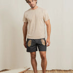 Man wearing a beige t-shirt and dark shorts with patterns, standing on a wooden floor with a white wall background.
