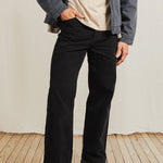 Person wearing black pants and a gray jacket on a wooden floor with a white wall background