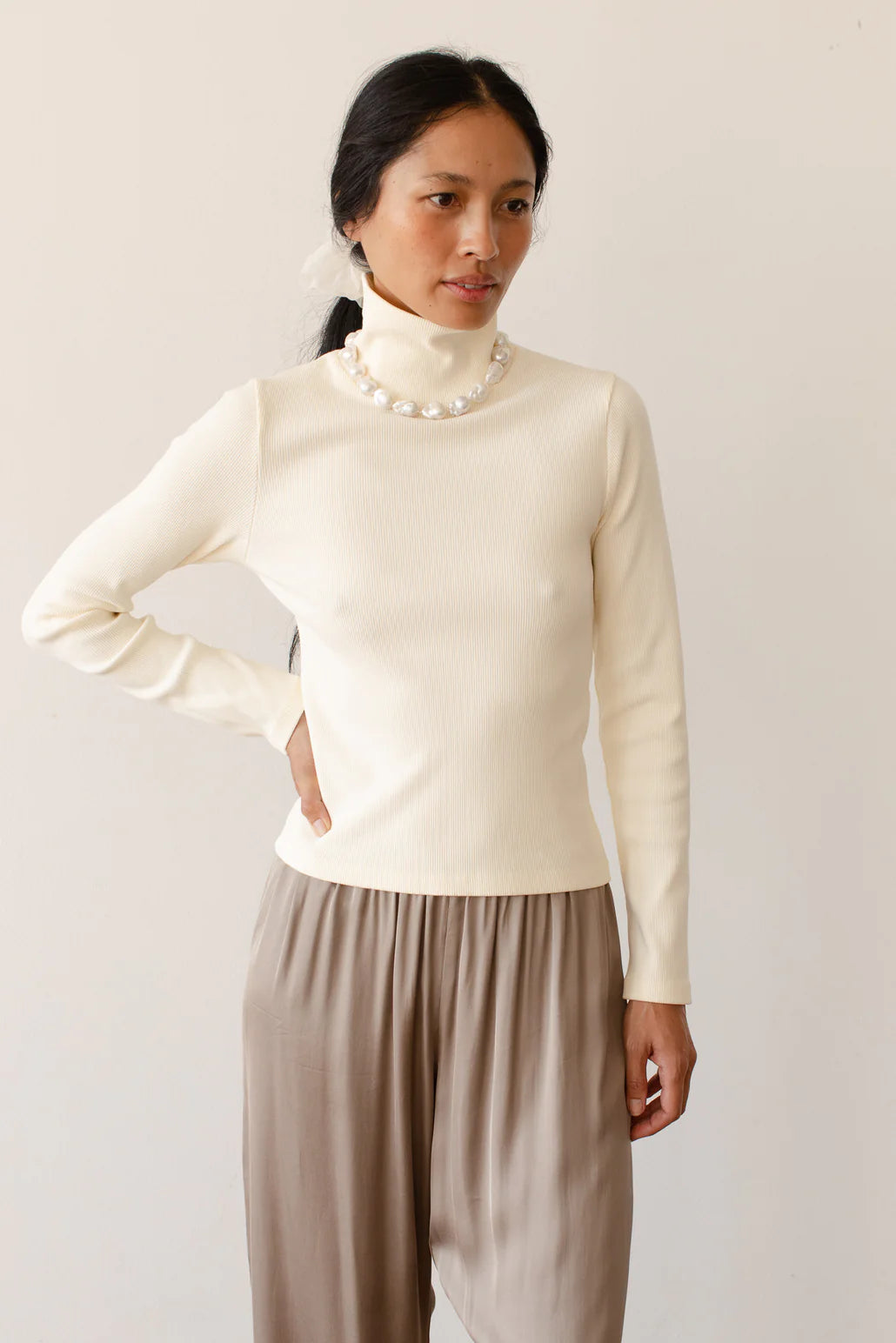 A medium shot image of a female model posing with a hand on her hip while wearing the Rib Turtleneck in Cream styled with a taupe trouser and pearl necklace