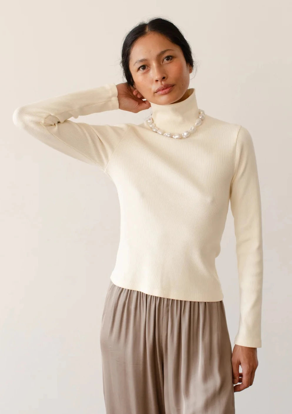 A medium shot image of a female model posing with an arm up to her head while wearing the Rib Turtleneck in Cream styled with a taupe trouser and pearl necklace