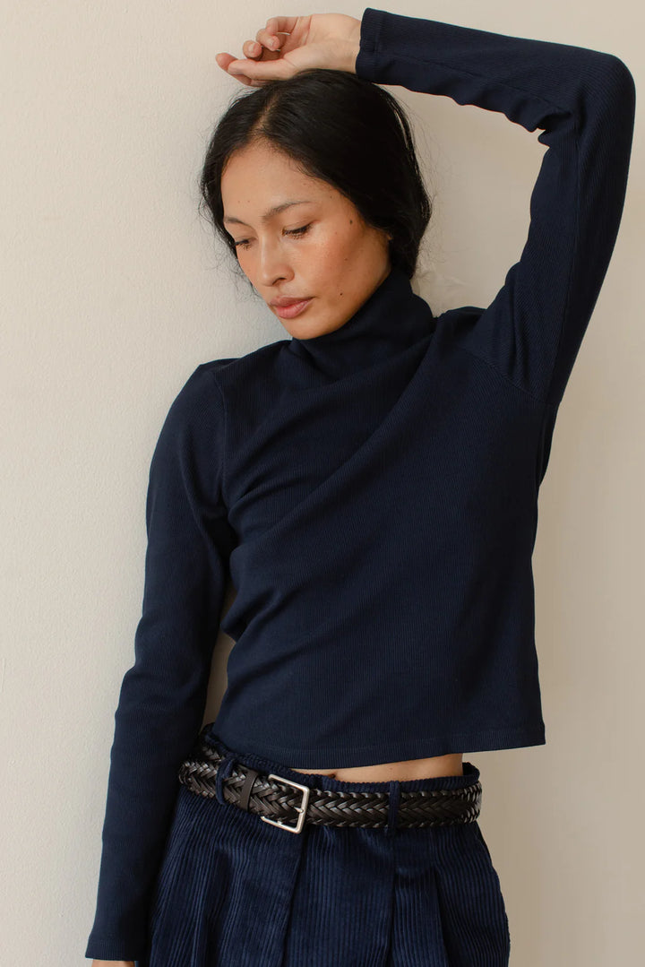 a medium close up image of a female model posing with her arm over her head while wearing the Rib Turtleneck in navy