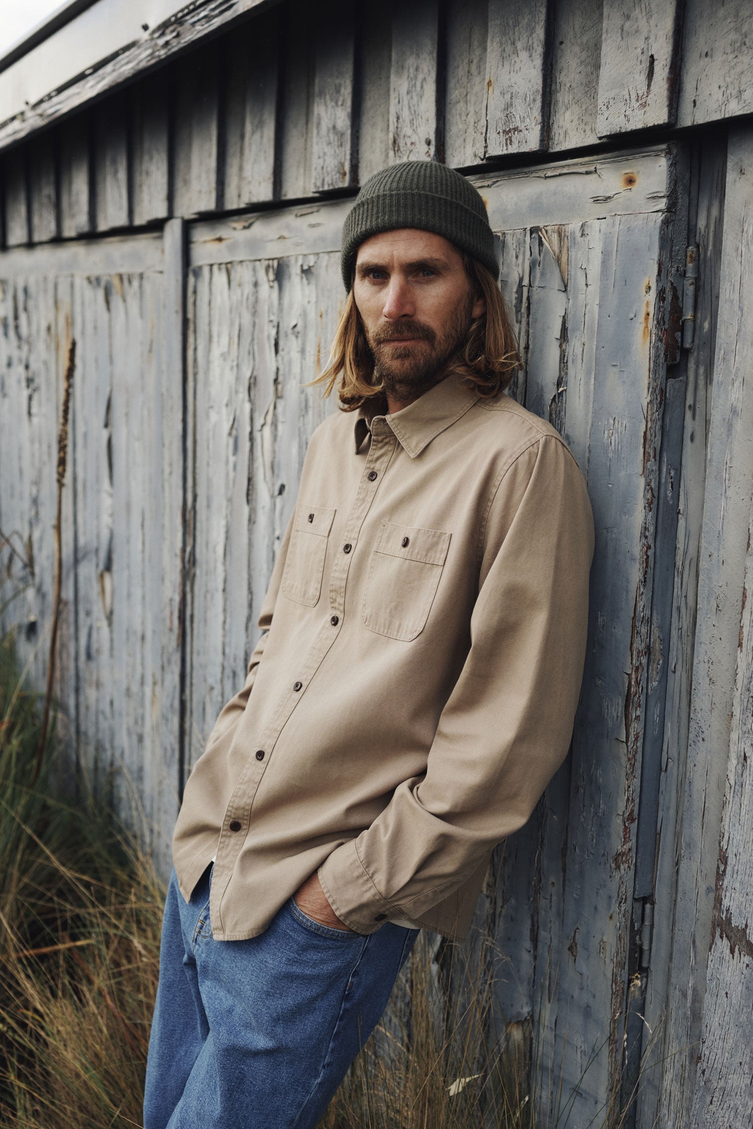 Man wearing a beige shirt and blue jeans leaning against a rustic wooden wall.