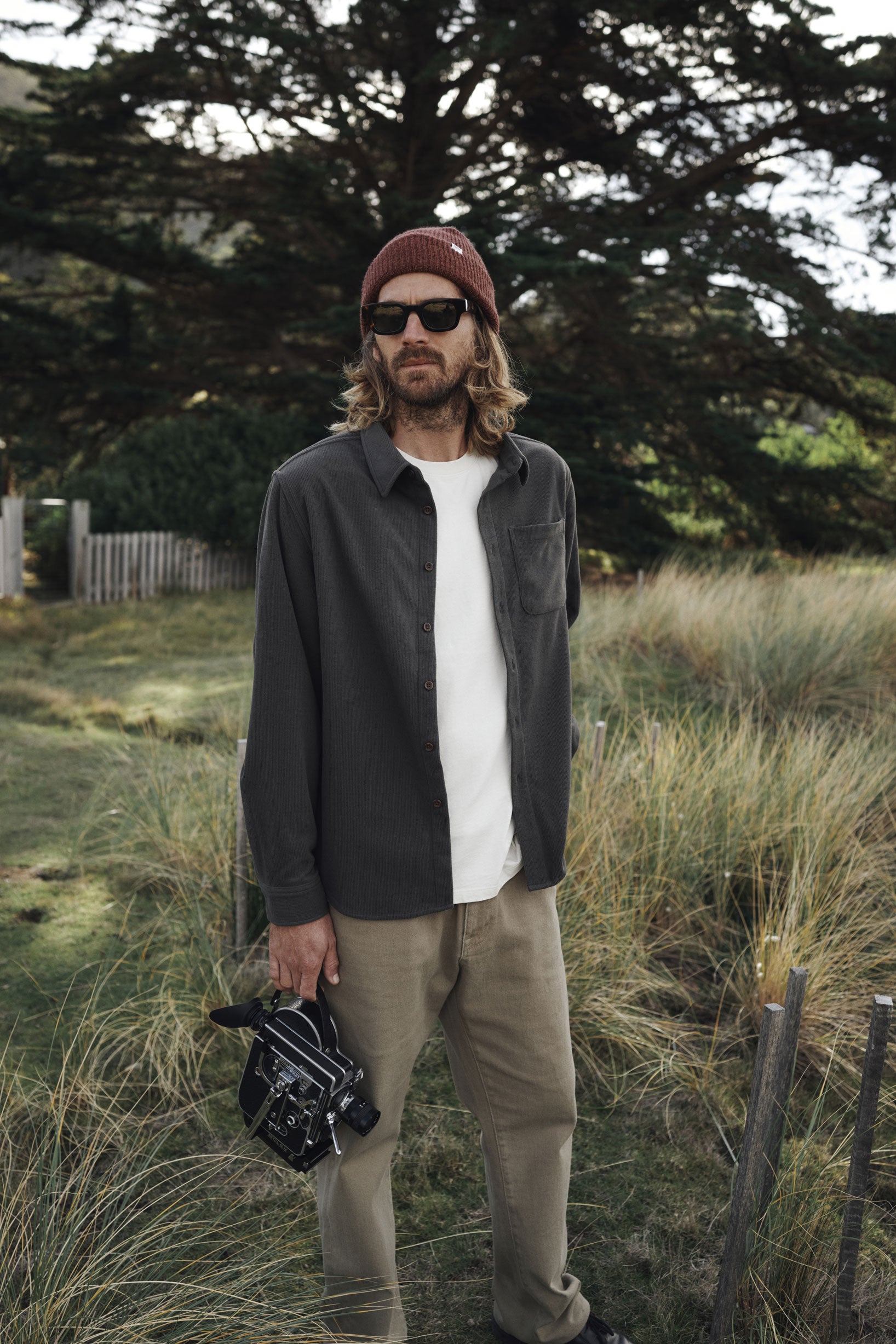 Man standing outdoors in a natural setting wearing sunglasses, a beanie, and casual clothing.