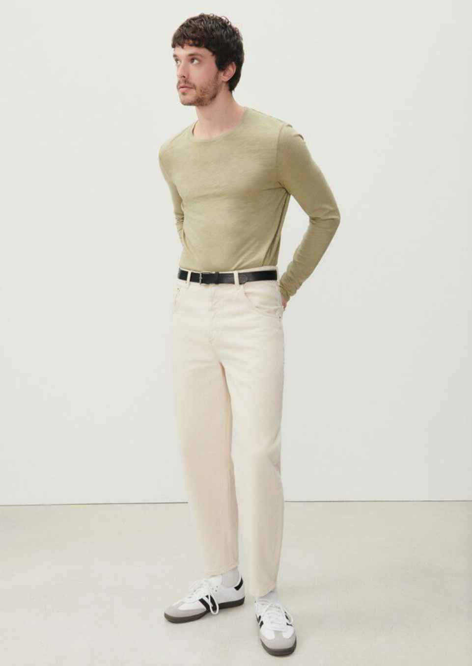 A full body image of a male model standing with both hands in his back pockets, wearing the Big Carrot Jeans in Ecru styled with a soft green long sleeve tucked in. 