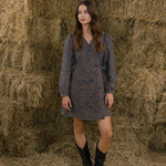 Woman in a patterned dress and black boots standing in front of hay bales.