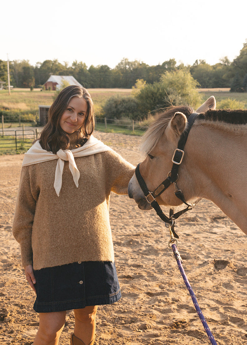Woman standing next to a horse in an outdoor setting with trees and fields in the background.