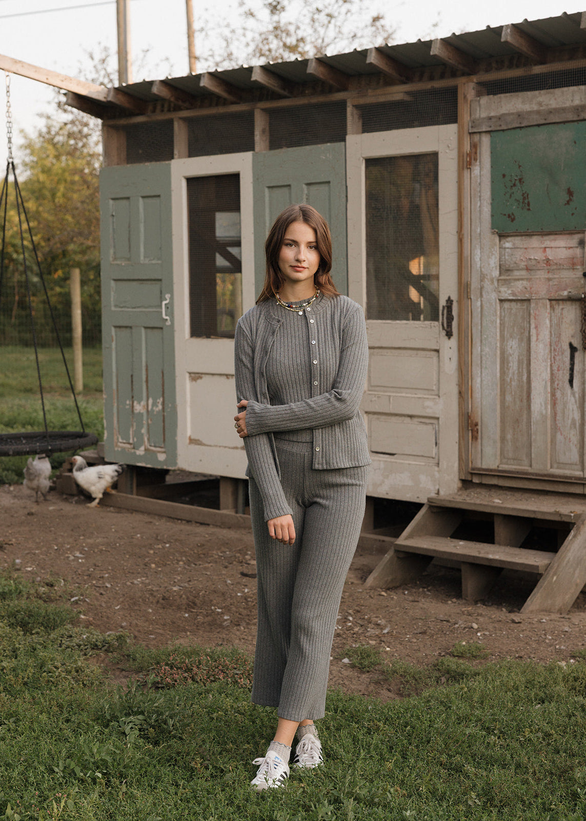Woman in a gray outfit standing in front of a rustic wooden shed with chickens in the background.