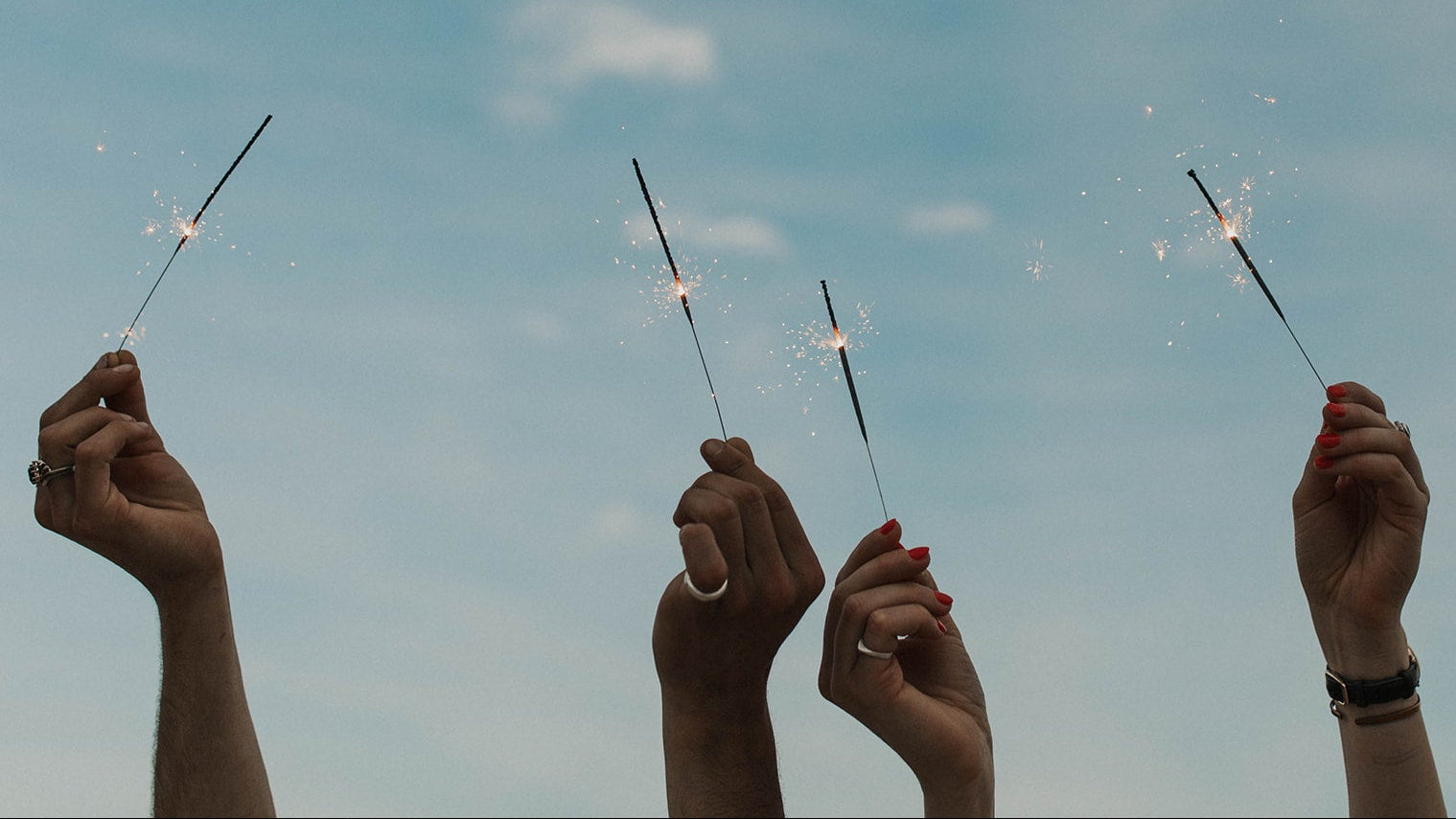 Four hands holding sparklers against a blue sky with scattered clouds.