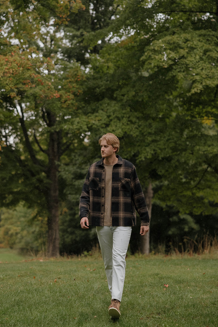 A male model is standing in the grass in front of a tree line while wearing the Joybird Carrot Jeans styled with a black and tan plaid shacket