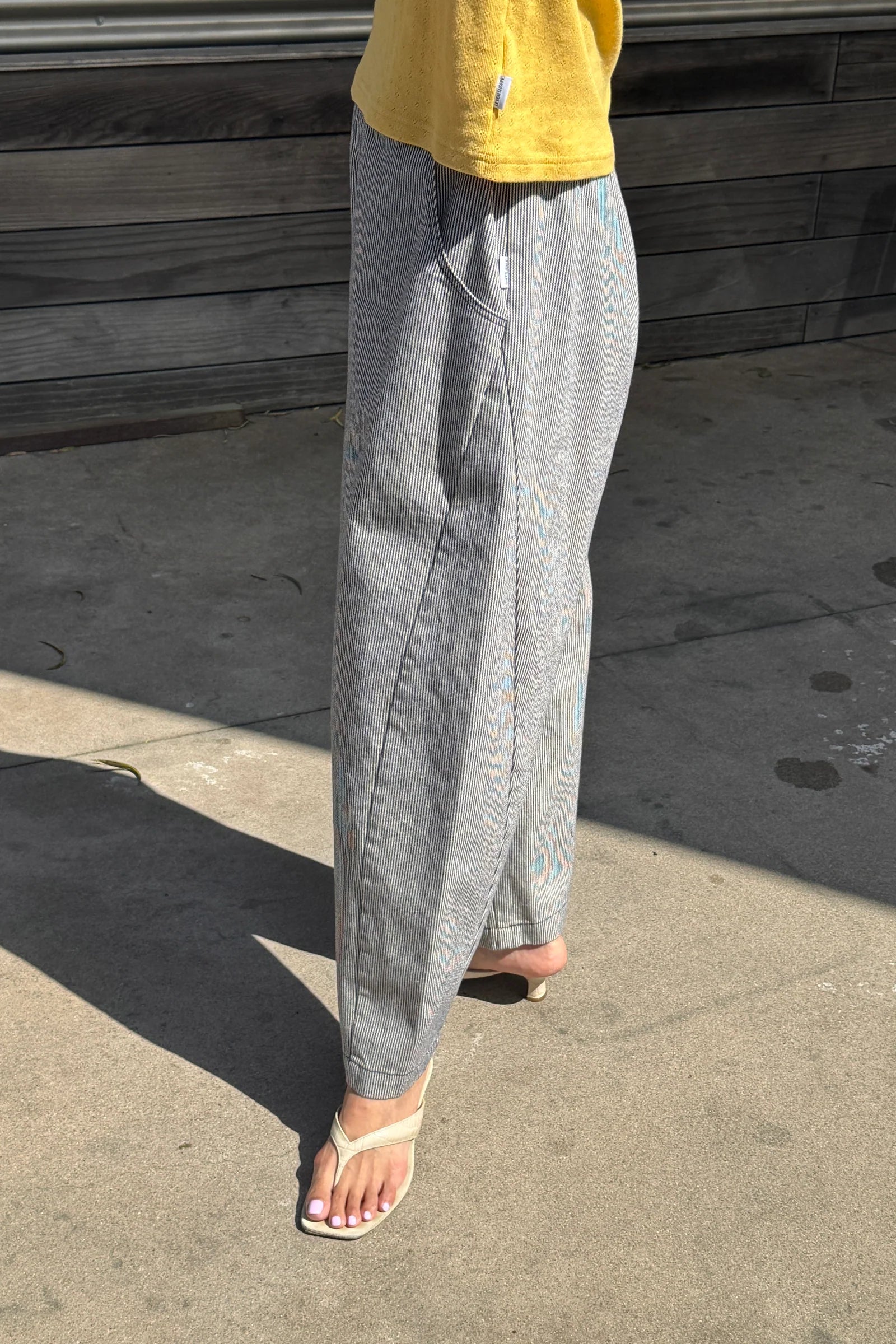 Person wearing white and blue striped pants and a yellow top standing on a concrete surface with wooden planks in the background.