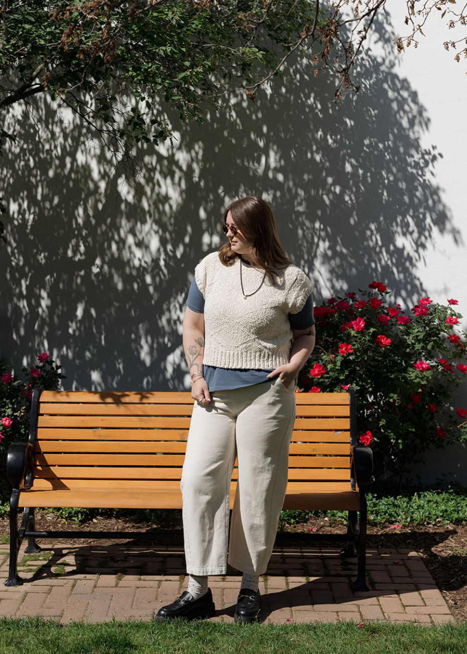 Woman standing next to a wooden bench in a garden with flowers and a white wall in the background