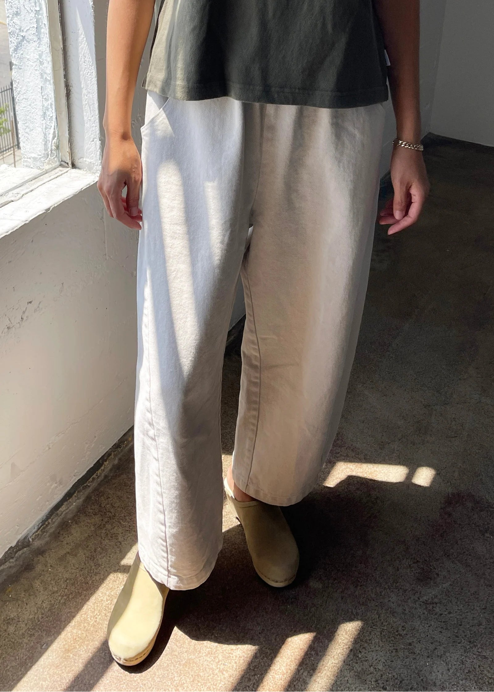 Person wearing a dark top and light-colored pants standing in a room with sunlight streaming in.