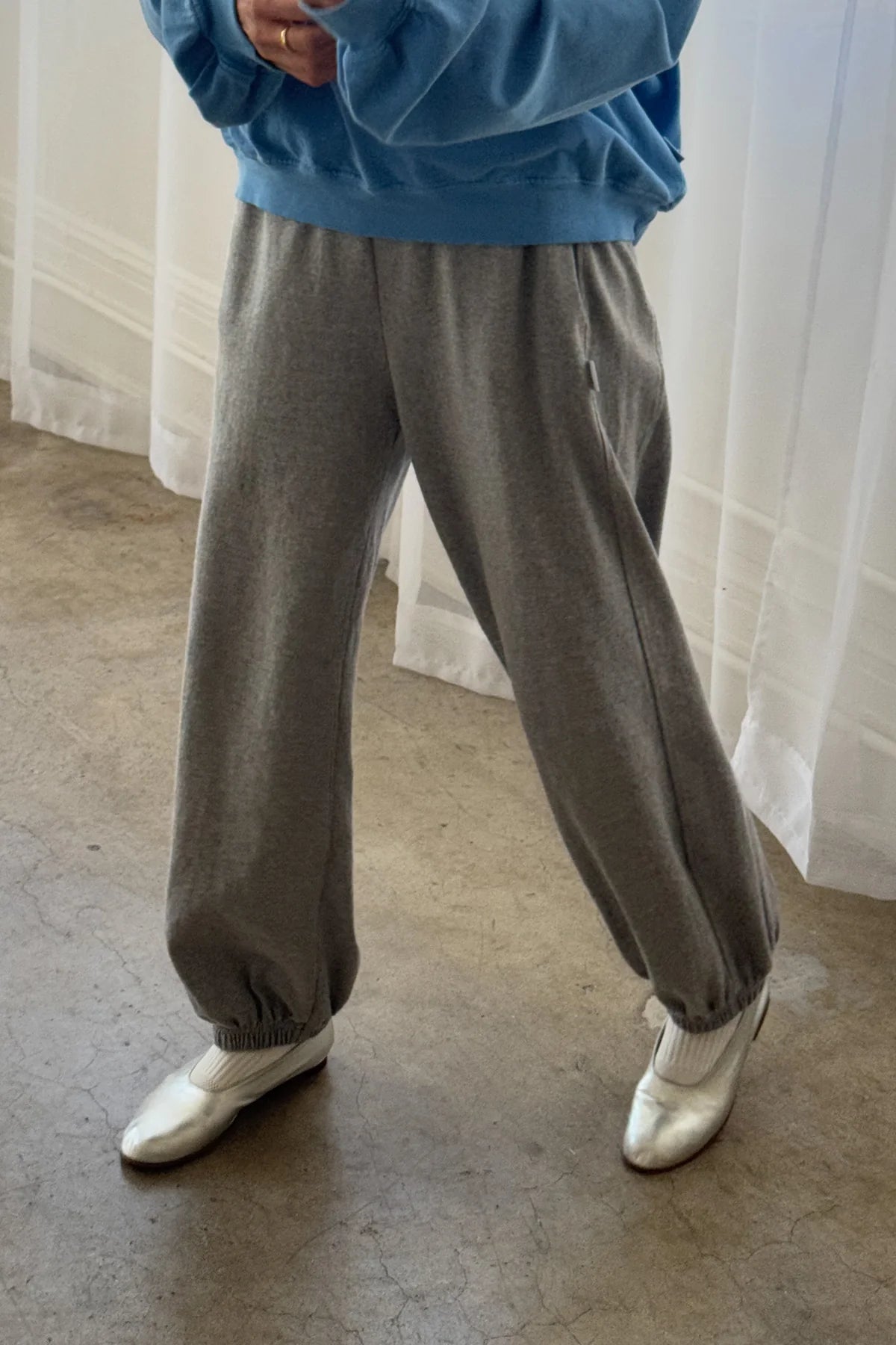 Person wearing gray sweatpants and a blue sweater with white shoes in front of a white curtain.