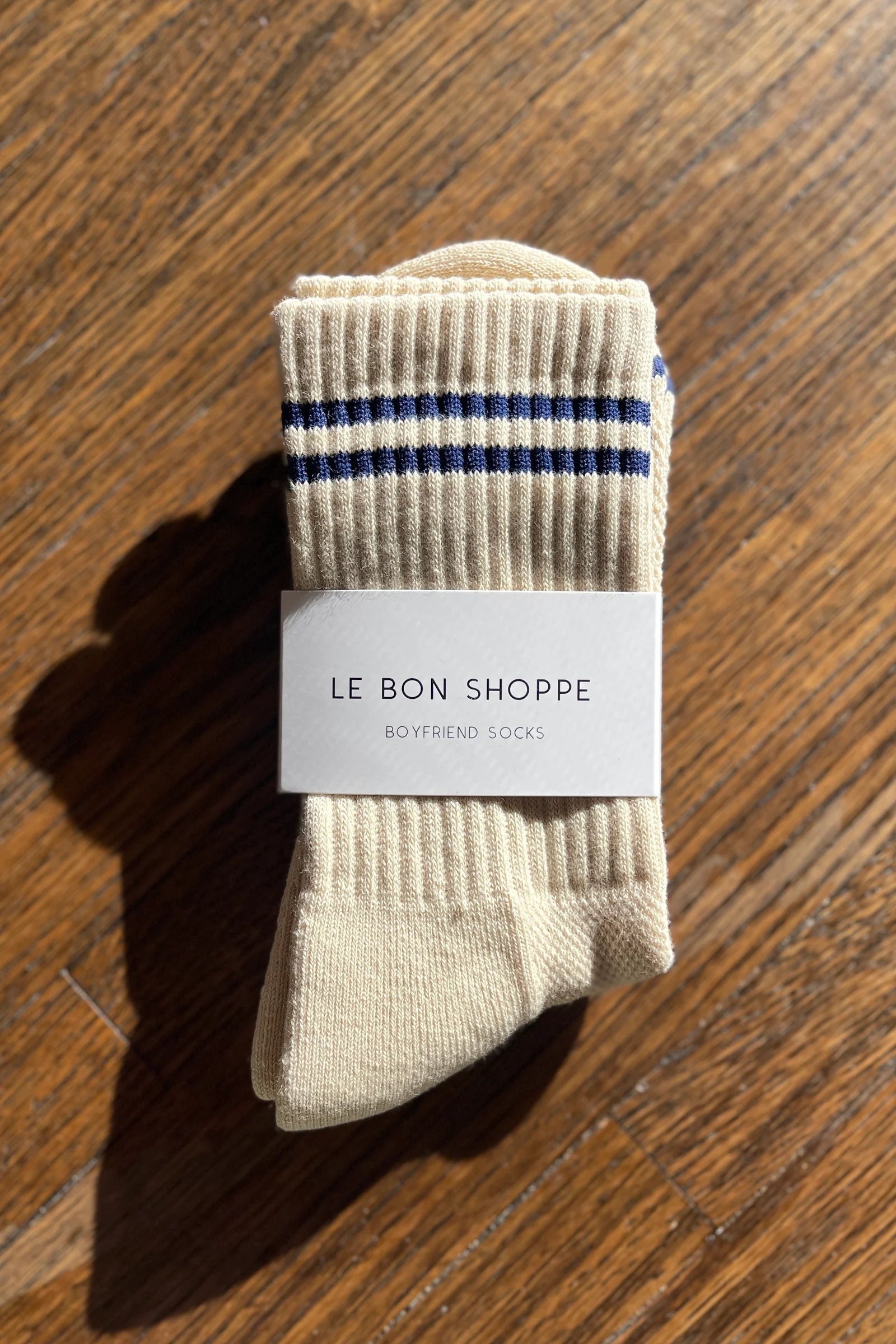 Pair of beige socks with blue stripes on a wooden surface, featuring 'Le Bon Shoppe' label.