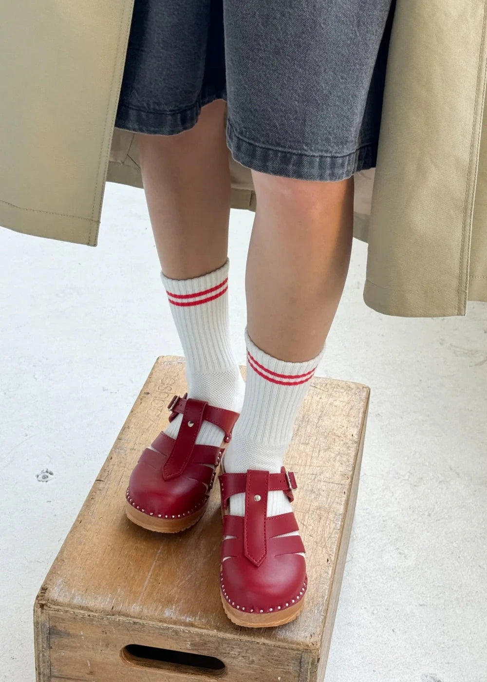 A person wearing clean white socks with a red double stripe and red closed-toe shoes standing on a wooden crate.