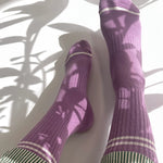 Purple socks with white stripes worn by a person on a white floor with plant shadows.