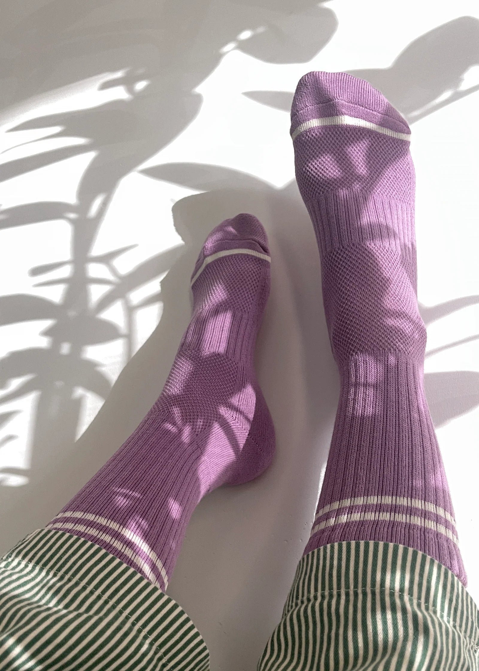 Purple socks with white stripes worn by a person on a white floor with plant shadows.