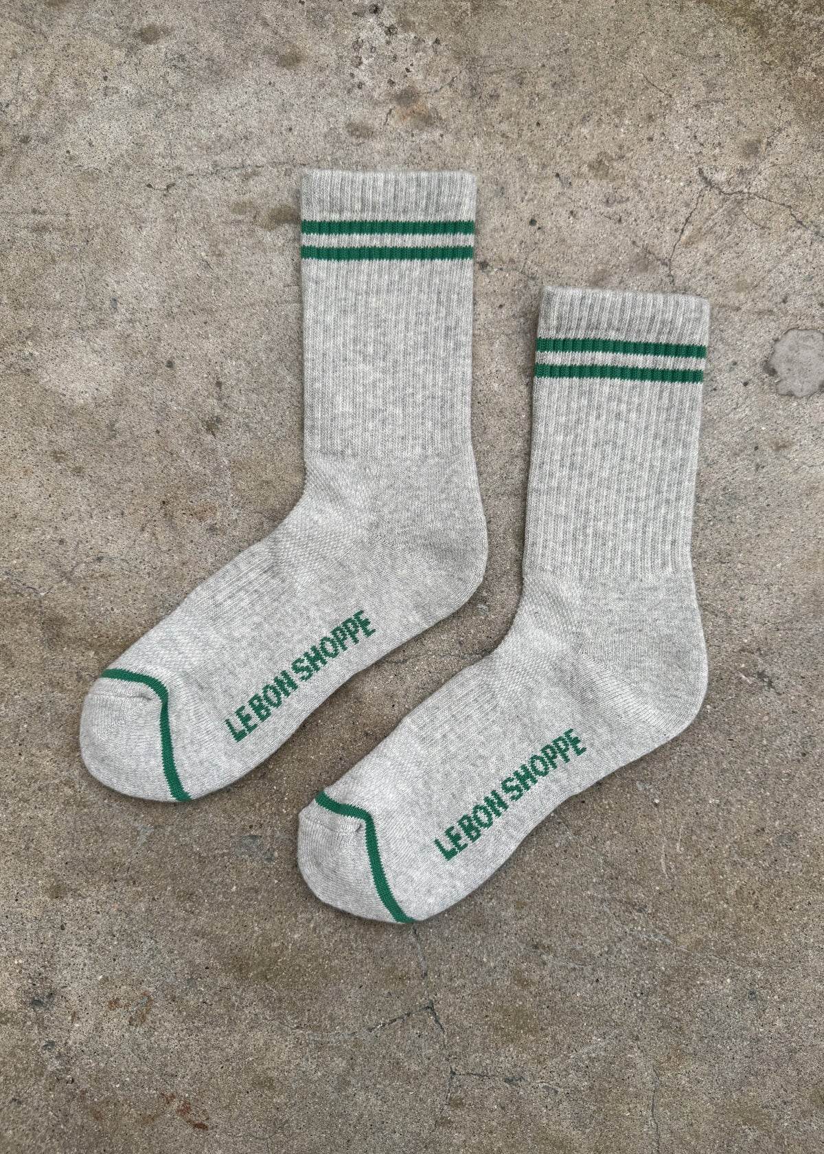 Gray socks with green stripes and 'Le Bon Shoppe' branding on a concrete surface