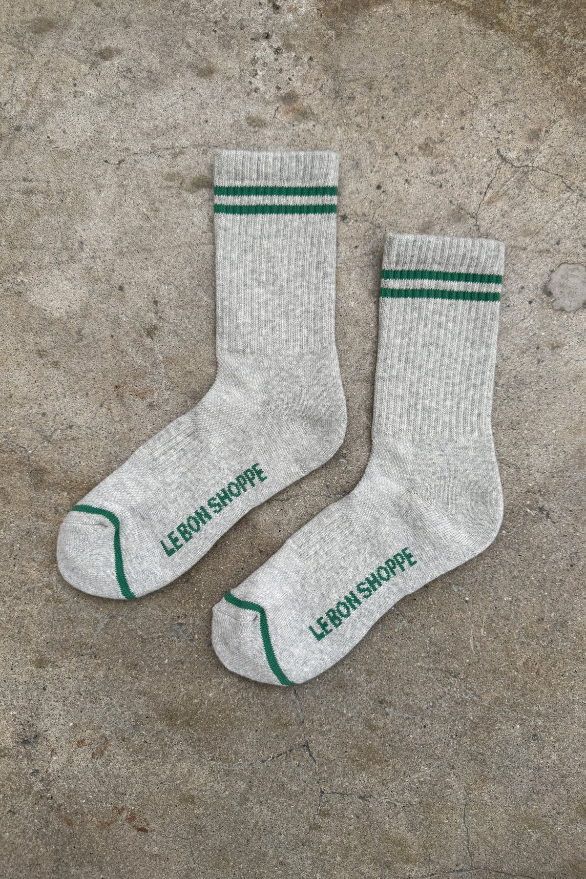 Gray socks with green stripes and 'Le Bon Shoppe' branding on a concrete surface