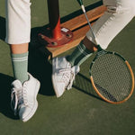 Person wearing white sneakers and green socks, holding a tennis racket on a tennis court.