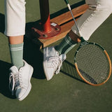 Person wearing white sneakers and green socks, holding a tennis racket on a tennis court.