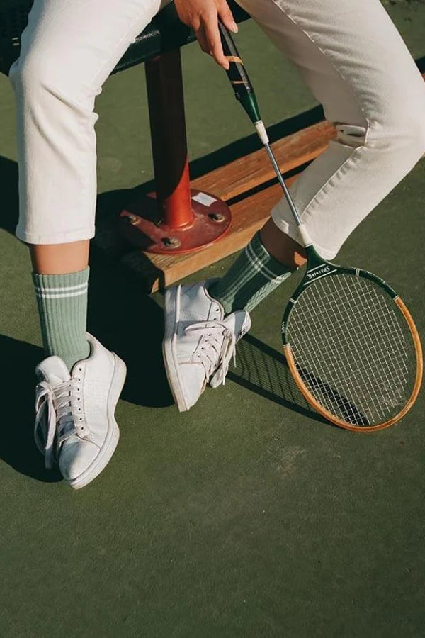 Person wearing white sneakers and green socks, holding a tennis racket on a tennis court.