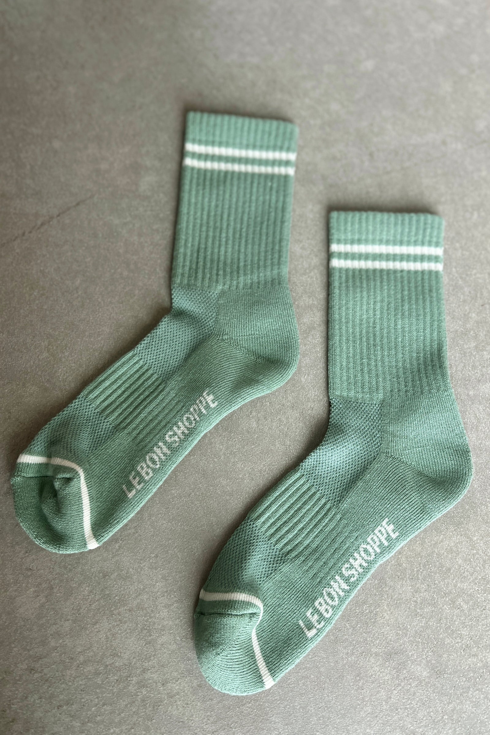 Two green socks with white stripes and 'Le Bon Shoppe' branding on a gray surface.