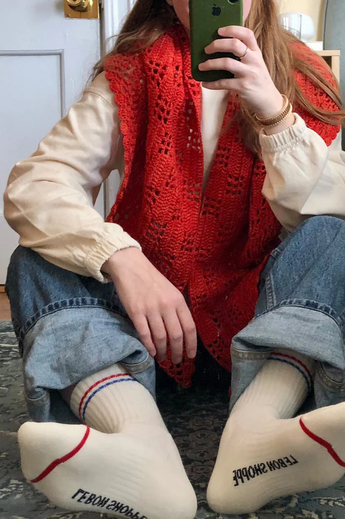 Person wearing a red knitted vest and white socks with text, sitting on a carpeted floor.
