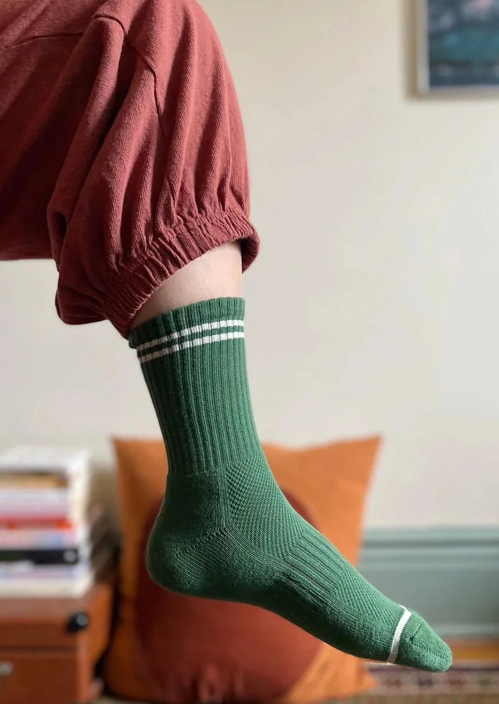 Person wearing green socks with white stripes and pink pants in front of a white wall and orange pillow.