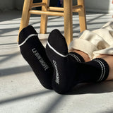 Person wearing black socks with 'Le Bon Shoppe' branding, sitting on a wooden stool.