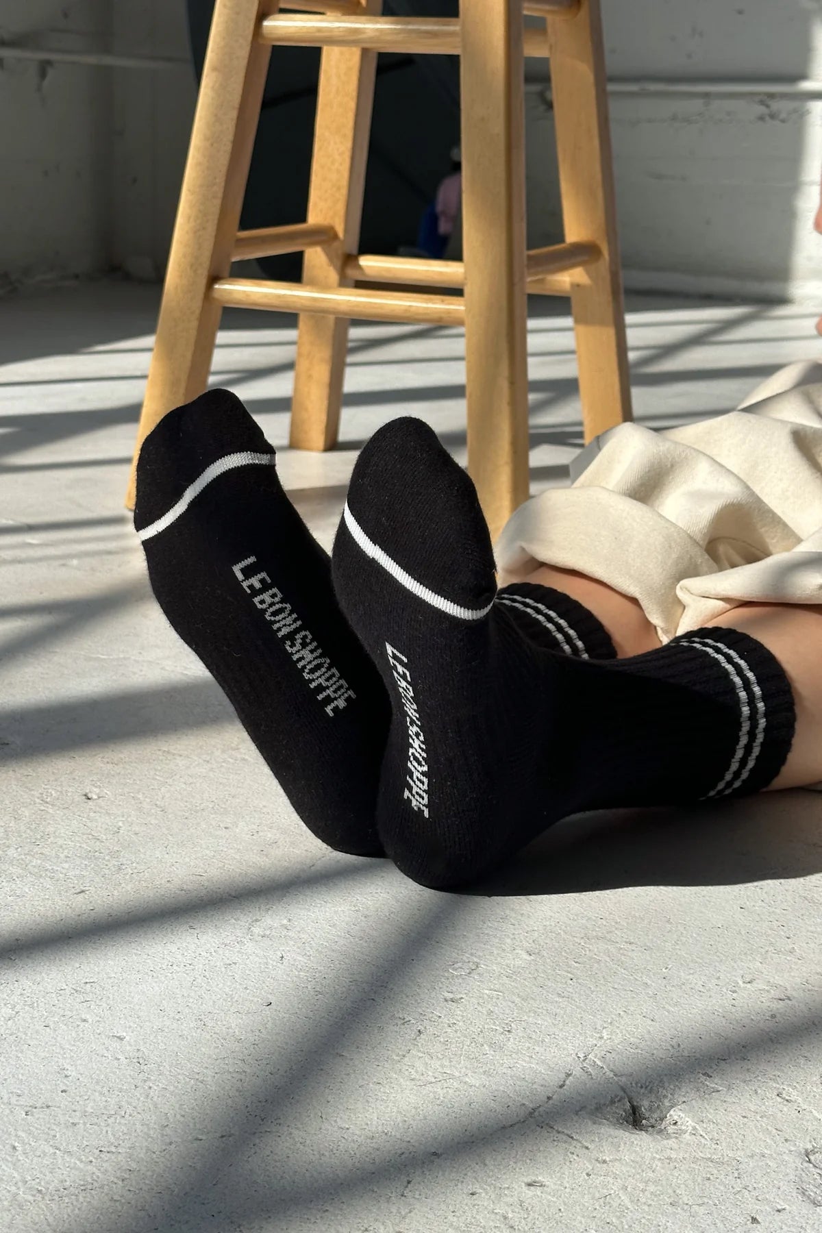Person wearing black socks with 'Le Bon Shoppe' branding, sitting on a wooden stool.