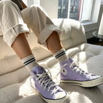 A person seated with oatmeal-colored boyfriend socks and white sneakers. The socks have a contrast double stripe and the shoes are converse style.