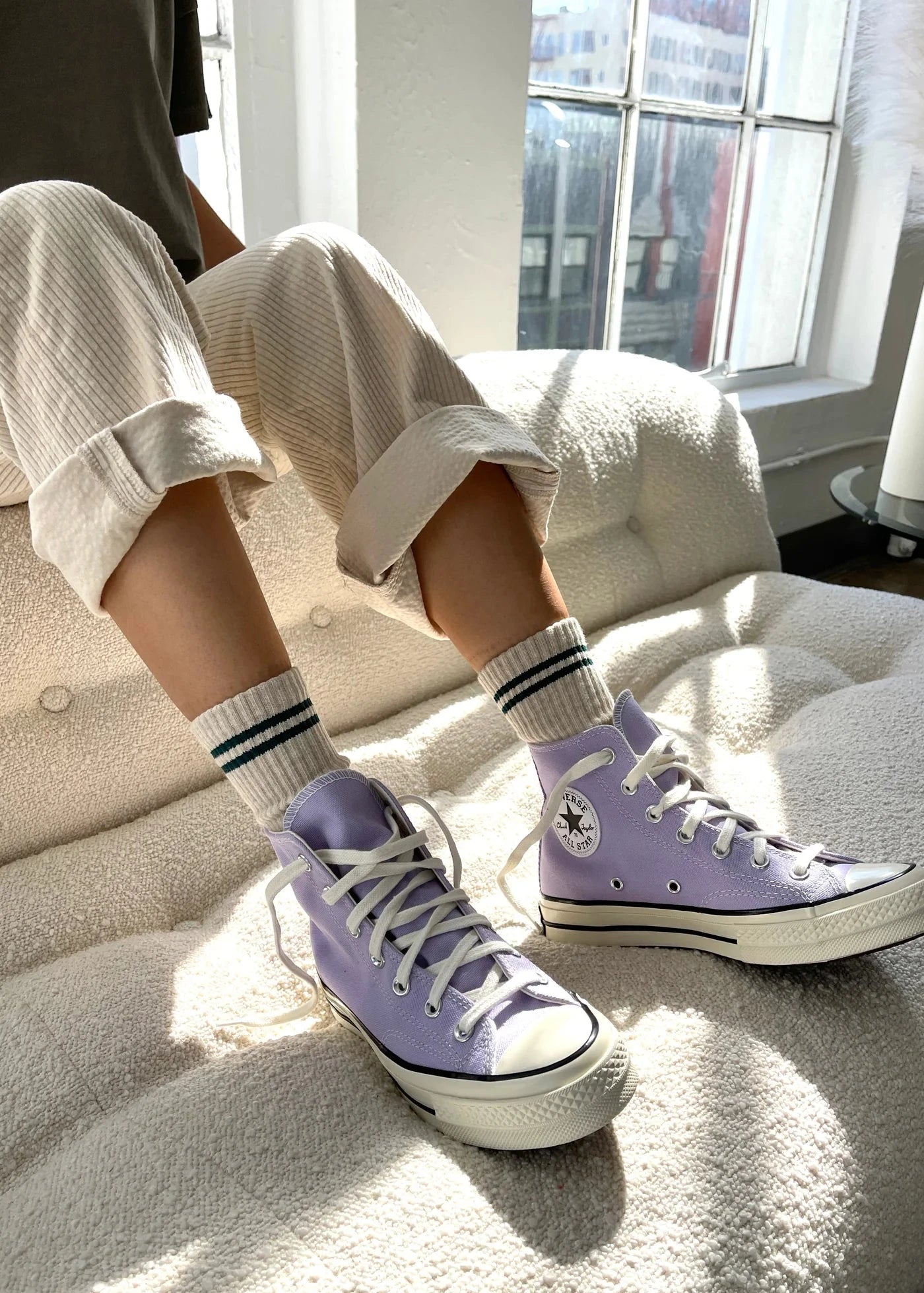 A person seated with oatmeal-colored boyfriend socks and white sneakers. The socks have a contrast double stripe and the shoes are converse style.