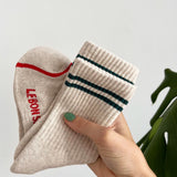 A hang holding tan socks with green stripes. The hand has painted green nails and is in front of a plant.