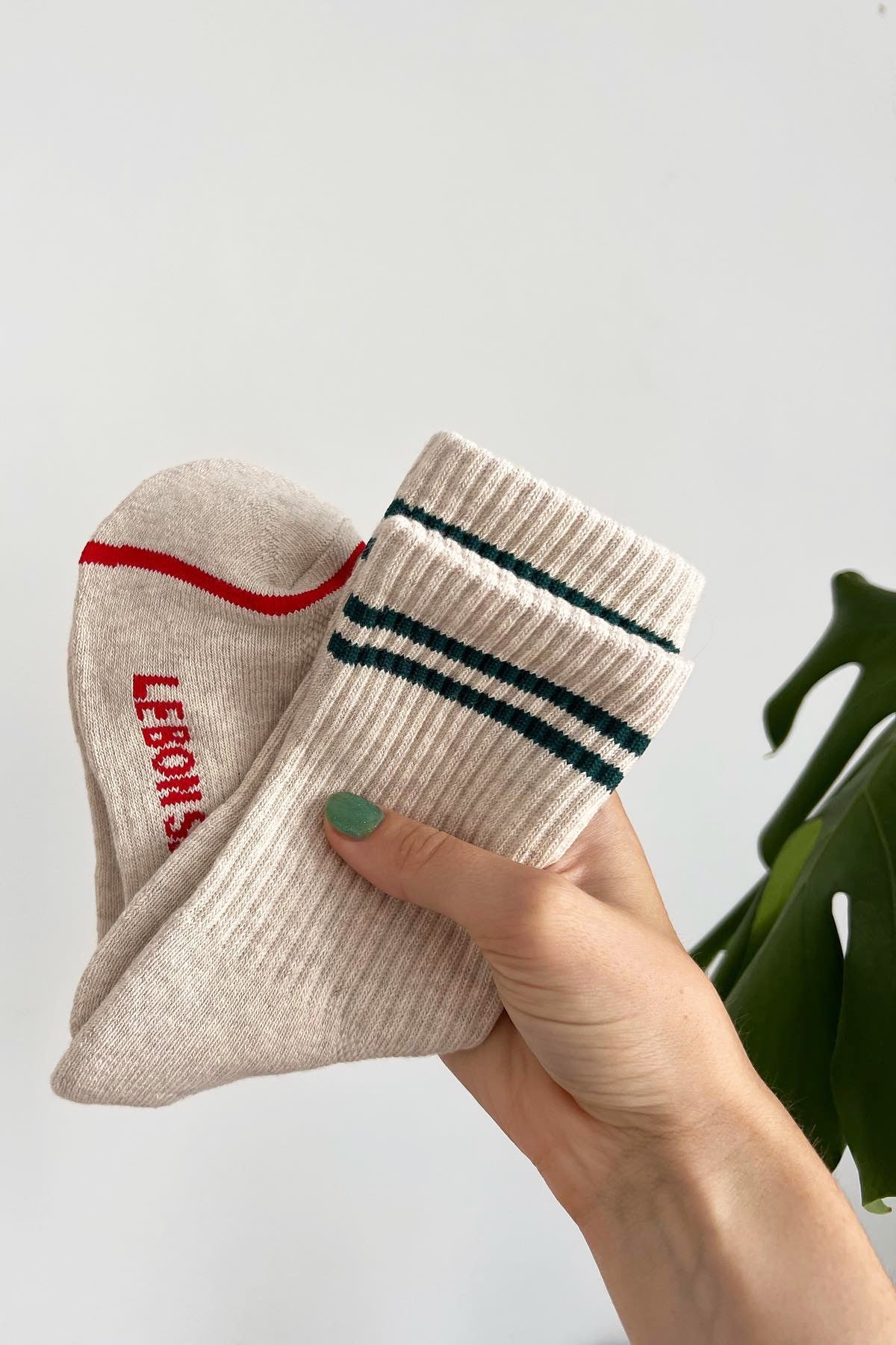 A hang holding tan socks with green stripes. The hand has painted green nails and is in front of a plant.
