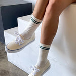 A pair of beige socks with a contrasting green double stripe on the cuffs, worn with white sneakers.