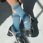 A person wearing powder blue socks with a contrast double stripe, paired with metallic-looking shoes, seated with a shadow falling over their lower body.
