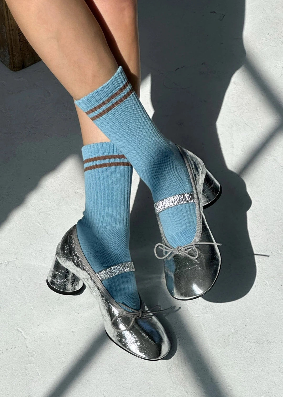 A person wearing powder blue socks with a contrast double stripe, paired with metallic-looking shoes, seated with a shadow falling over their lower body.