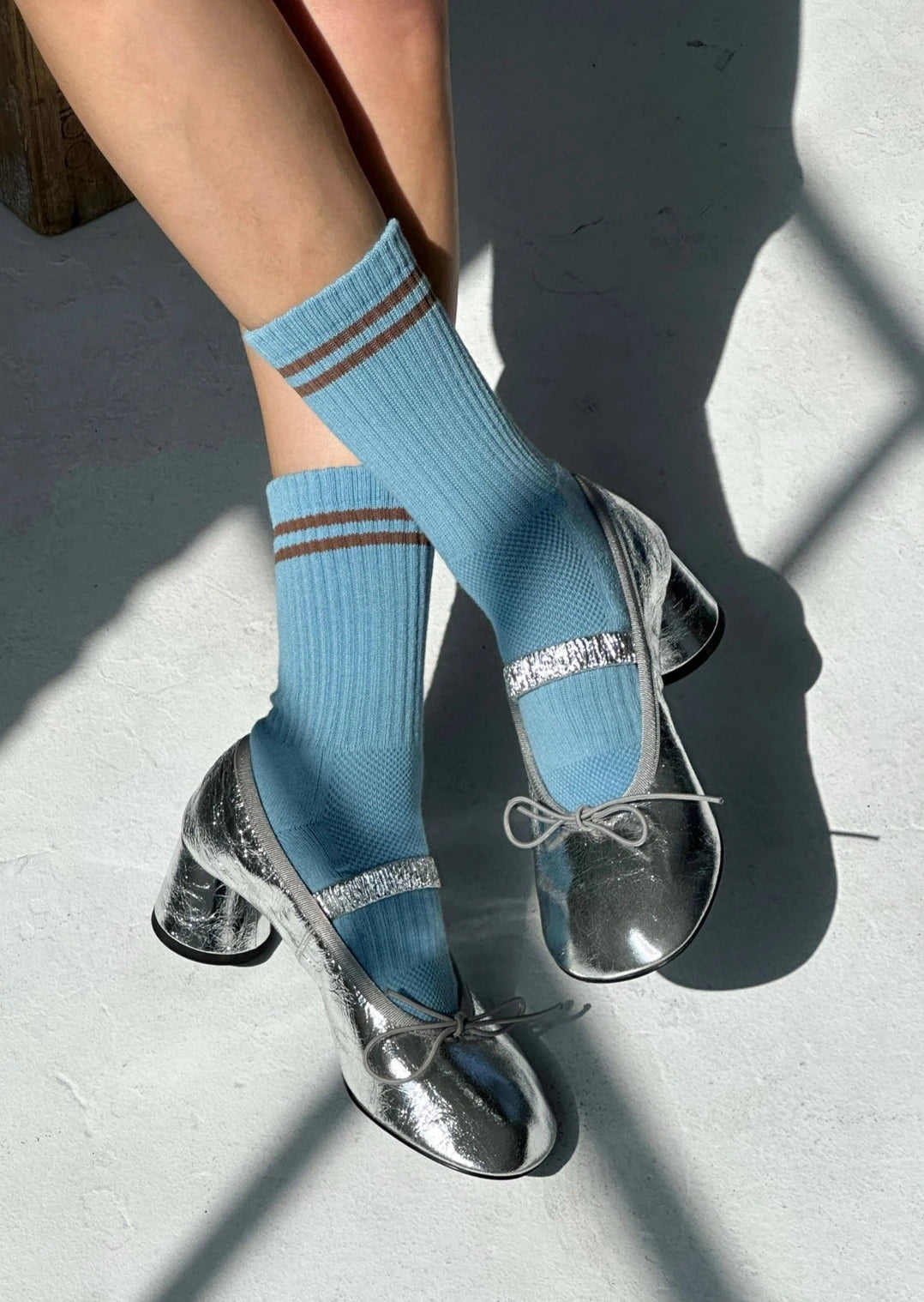 A person wearing powder blue socks with a contrast double stripe, paired with metallic-looking shoes, seated with a shadow falling over their lower body.