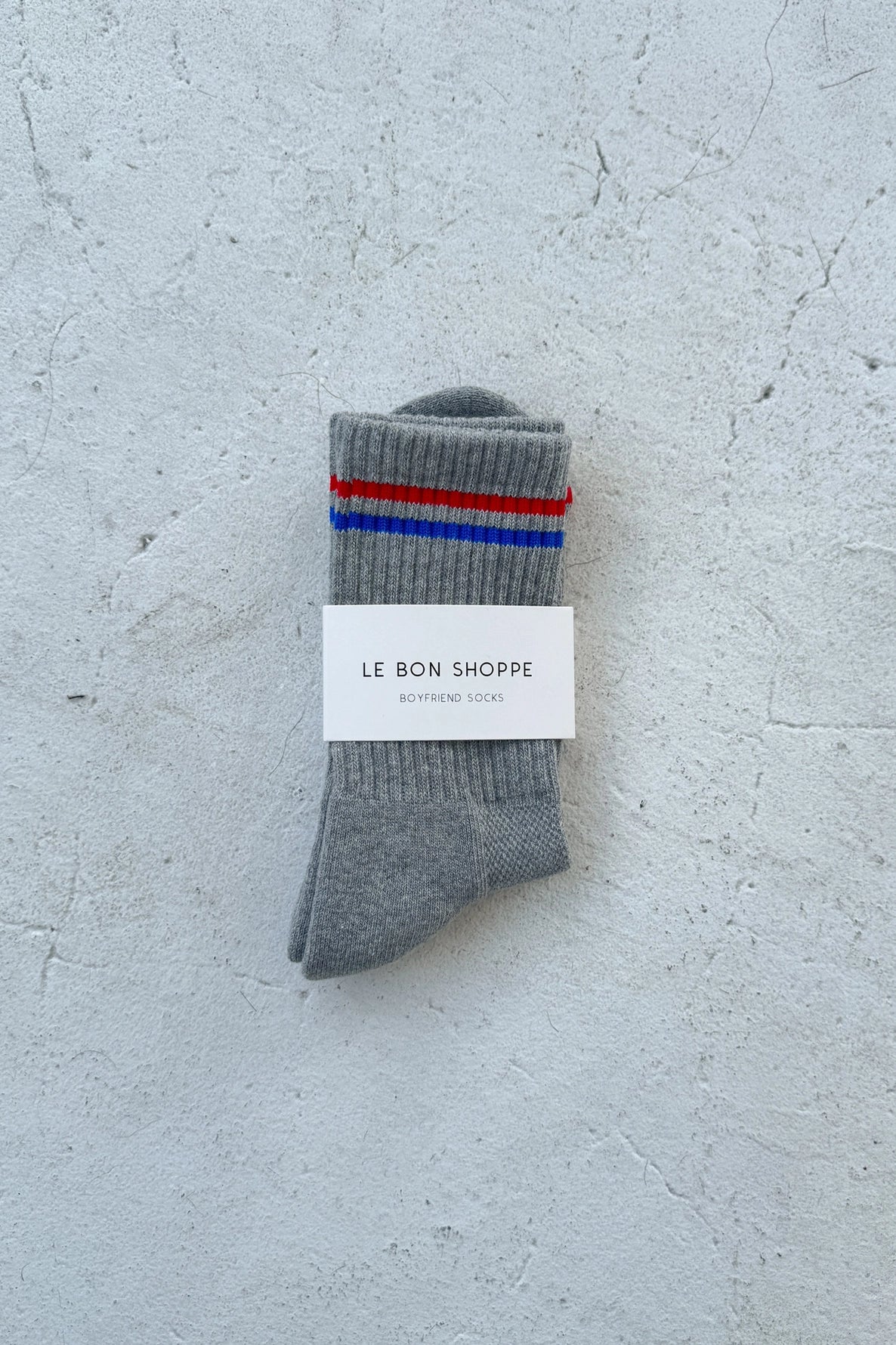 Grey socks with red and blue stripes and Le Bon Shoppe branding on a white background.