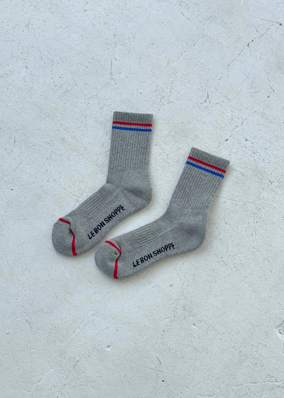 Grey socks with red and blue stripes and Le Bon Shoppe branding on a white background.