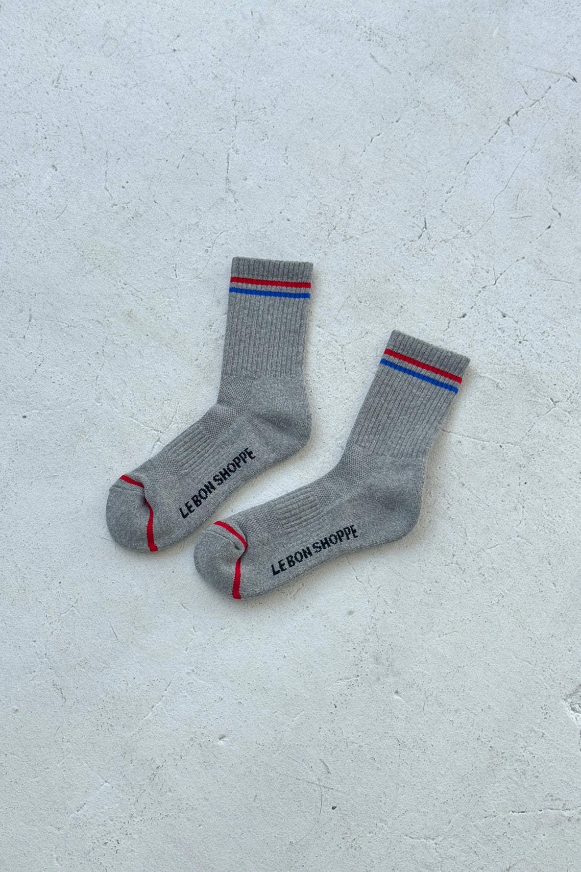 Grey socks with red and blue stripes and Le Bon Shoppe branding on a white background.