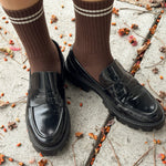 Brown knee-high socks with white stripes worn with black shoes on a concrete surface with scattered leaves.