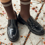 Brown knee-high socks with white stripes worn with black shoes on a concrete surface with scattered leaves.