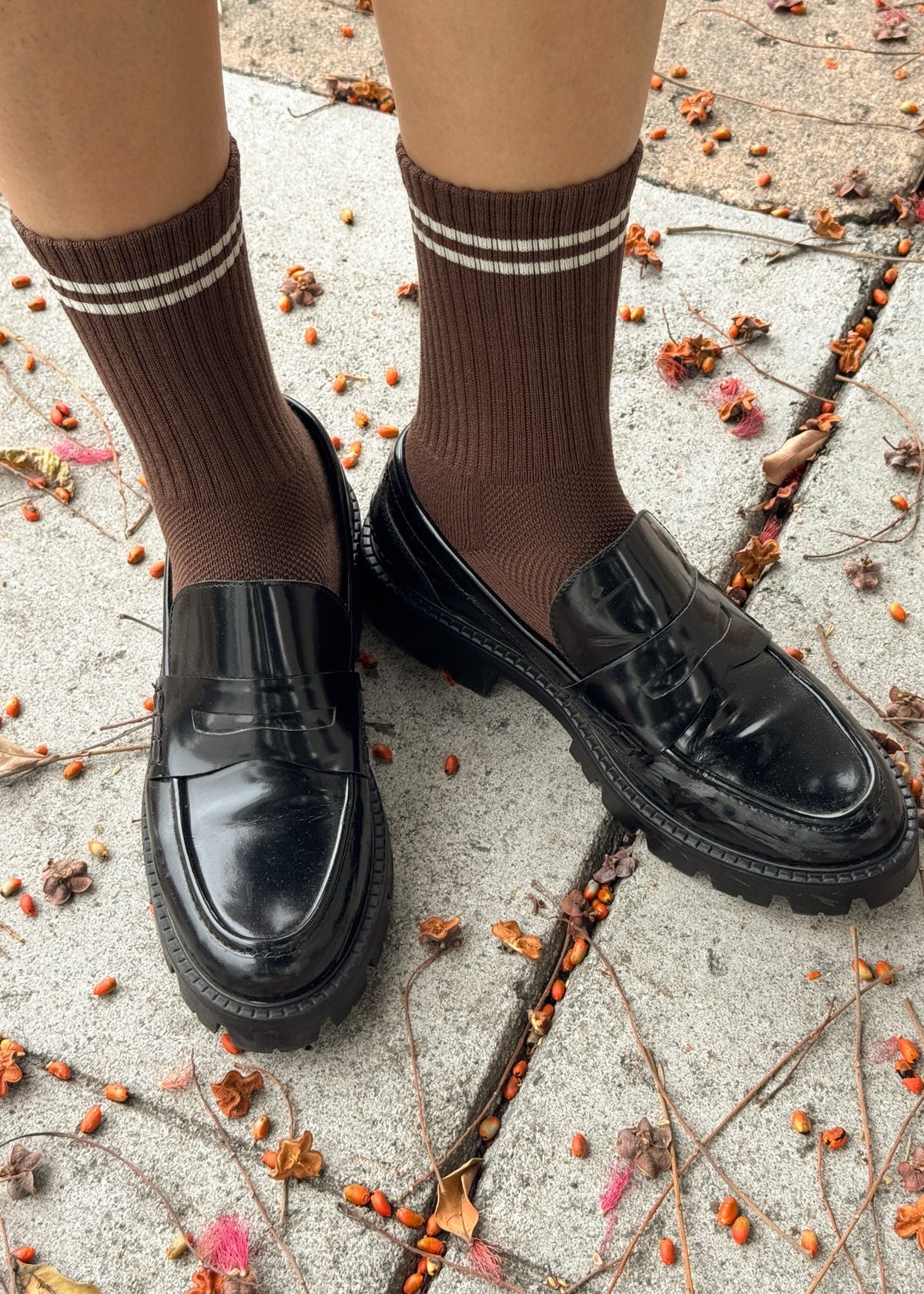 Brown knee-high socks with white stripes worn with black shoes on a concrete surface with scattered leaves.