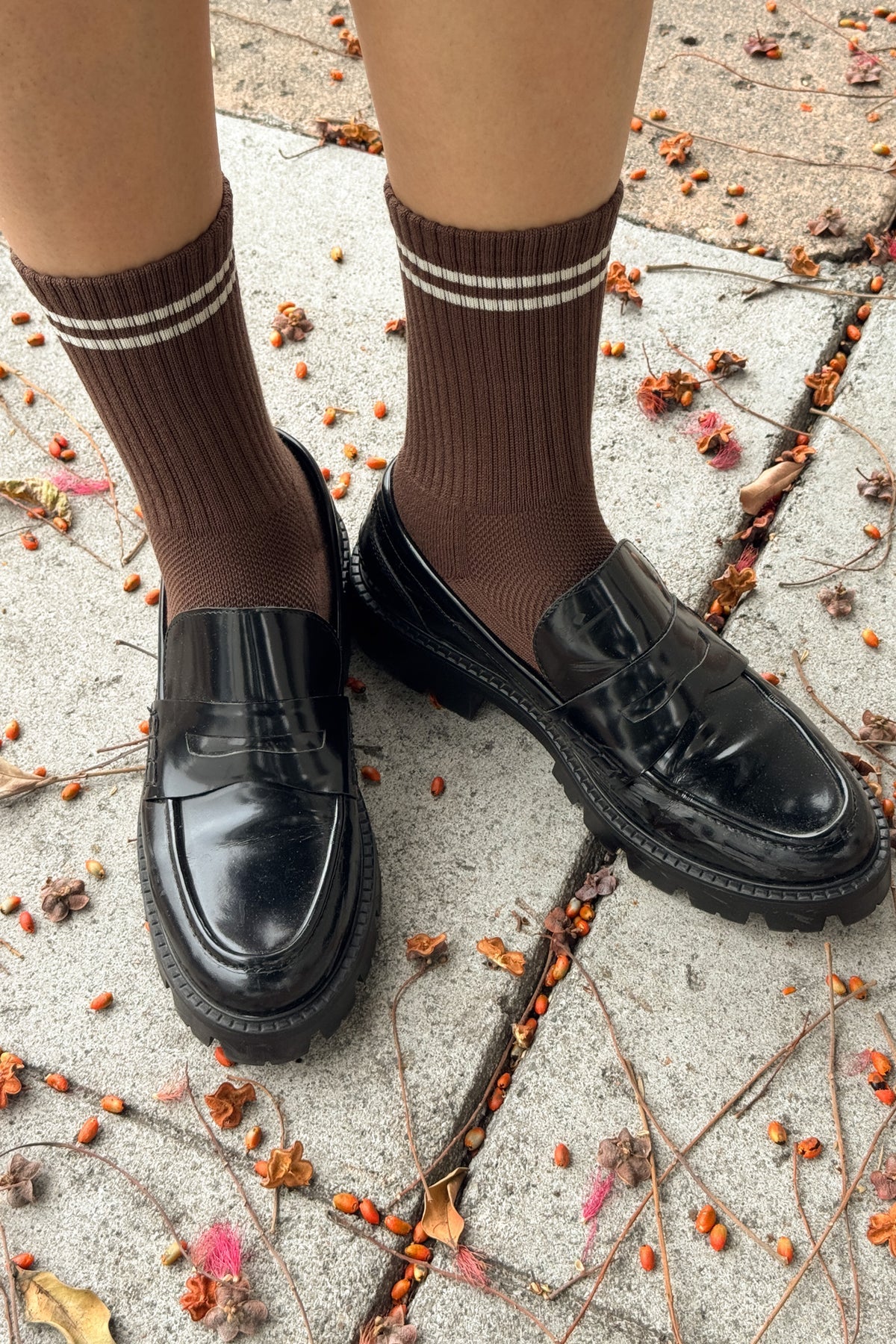 Brown knee-high socks with white stripes worn with black shoes on a concrete surface with scattered leaves.