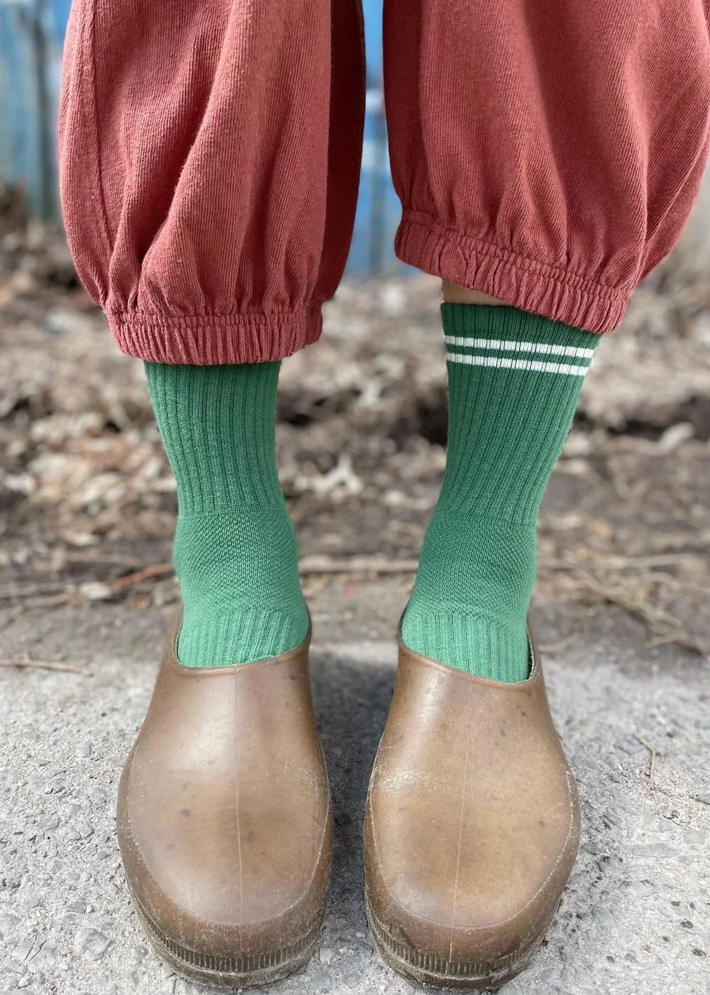 Person wearing garden shoes, green socks with white stripes and pink pants standing on a sidewalk.