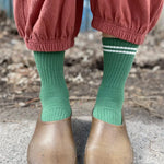 Person wearing garden shoes, green socks with white stripes and pink pants standing on a sidewalk.